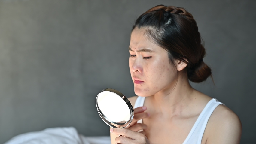 Worried Asian woman with bad emotional while looking acne and scar occur on her face by mini mirror. Conceptual shot of Acne and Problem Skin on female face. | Shutterstock HD Video #1085502596