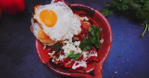 Top view of clay bowl of tasty red chilaquiles with fried egg on black surface. Vegetables, herbs, and delicious traditional dish with sauce and eggs from above. Authentic Mexican food
