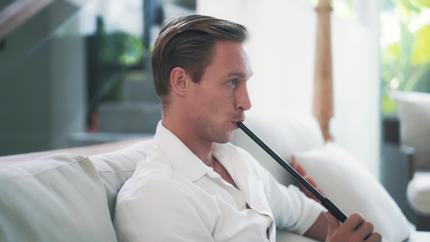 Tired young beautiful man sits on armchair and smoking hookah in slow motion. Pretty Caucasian guy lets out clouds of smoke and rests. Art of hookah smoking. High quality FullHD footage