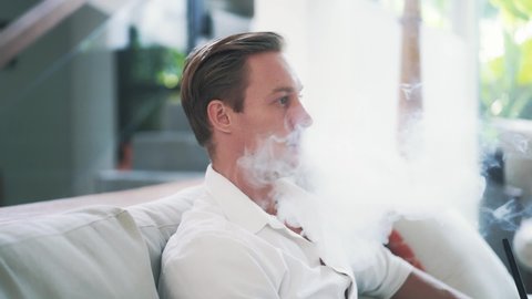 Tired young beautiful man sits on armchair and smoking hookah in slow motion. Pretty Caucasian guy lets out clouds of smoke and rests. Art of hookah smoking. High quality FullHD footage