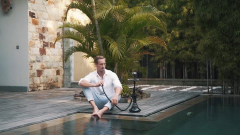 Young Caucasian man sits at the poolside and smoking hookah in slow motion. Successful young guy relax after hard day with hookah. The art of hookah smoking. High quality FullHD footage