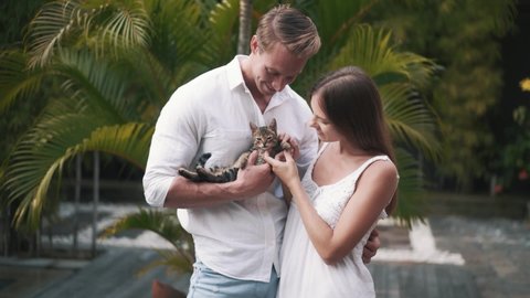Young Caucasian couple holds little kitten in hands and hugs in slow motion. Beautiful man and woman play with cute tabby kitten and hug. Romantic couple, animal lovers. High quality FullHD footage