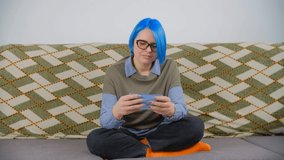 Millenial playing game on smartphone. White female gaming on modern mobile phone with smile. Individual person with bright blue hair using cellphone to play video games in leisure time