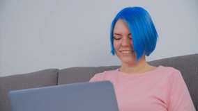 Happy white woman with blue hair working on laptop with cheerful toothy smile. Young creative professional doing freelance work on notebook at home during lockdown. Cute female typing on computer