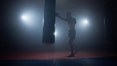 Sportsman boxing in smoky studio. Kickboxing. Silhouette of young man training with punching bag in boxing club. Male strikes with his feet. Darkness background. Sport concept. 4K, UHD