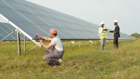 Indian technician examining solar panels with multimeter. Indian man in uniform squatting near solar panel with multimeter in hands. Two business people with gadgets talking on background