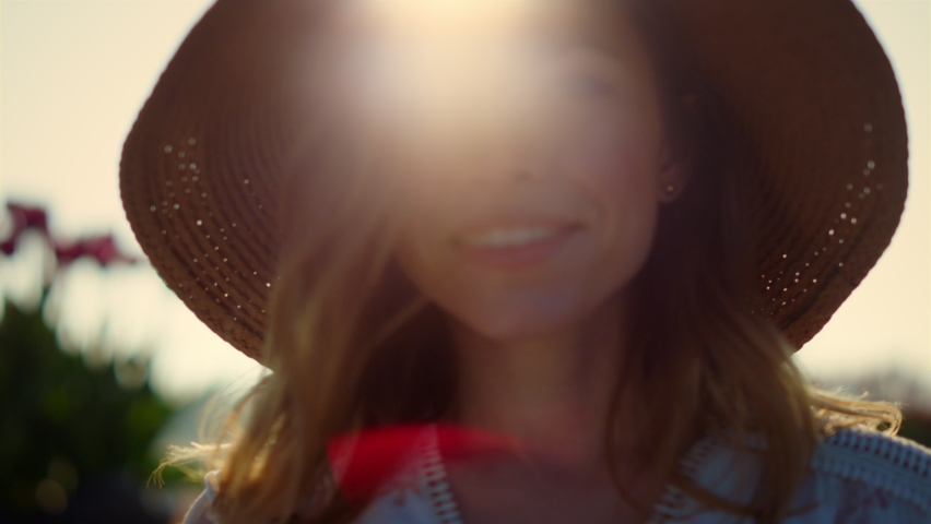 Beautiful woman in straw hat smiling to camera in sun reflection outdoors. Portrait of pretty girl in sunhat looking with smile in sunset light outside. Close up happy woman face in evening lights.