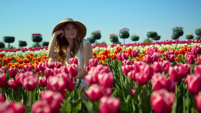 Relaxed young woman sitting in beautiful spring garden. View of colorful blooming tulip field. Romantic girl watching floral landscape in summer park. Pretty lady enjoying sunny day in flowers outside