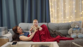 Happy mother playing with her toddler lying on the sofa, mom kisses hugs her 10 month old baby boy. Family with cat resting at home in the evening. High quality 4k footage