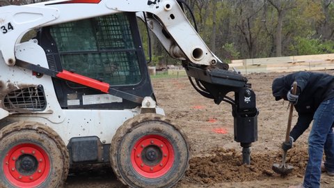 andover , United States - 01 15 2022: Construction worker uses shovel to remove loose dirt while skid steer loader with hydraulic auger removes auger from hole and drives forward to remove dirt from a