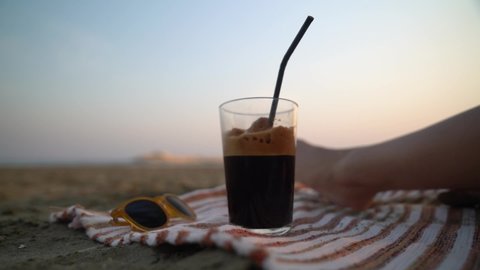 Iced coffee at the beach. Close-up as a woman grabs her iced coffee from the beach blanket with a relaxing sandy beach and ocean background.
