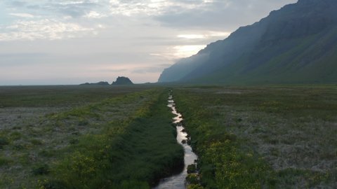 Backwards fly above small creek flowing through flat meadow. Steep rocky escarpment aside and sun behind clouds. Revealing standing SUV car. Iceland