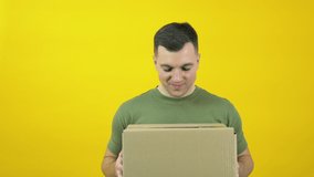 Young delivery man courier in a green T-shirt lifts a craft cardboard box in front of him. The guy is standing in front of a yellow background with a parcel in his hands. Yellow-green color scheme