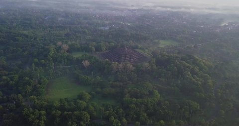 Reveal drone shot of Borobudur Temple in the morning and slightly foggy weather. It is surrounded by green tree vegetation. The Temple is empty from visitor