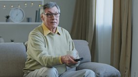 Grey-haired aged man in glasses sitting on couch watching TV enjoying relaxing at home drinking tea coffee aged male uses remote control to switch channels smiling enjoy funny movie television program