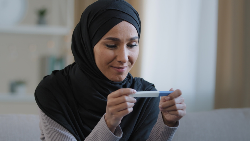Portrait joyful surprised girl in hijab holding pregnancy test sit in living room excited with positive result beautiful islamic young woman feel happy after successful fertility treatment IVF concept Royalty-Free Stock Footage #1085525420