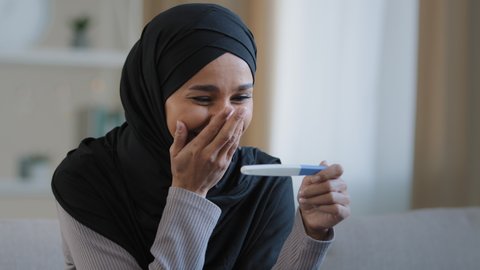 Portrait joyful surprised girl in hijab holding pregnancy test sit in living room excited with positive result beautiful islamic young woman feel happy after successful fertility treatment IVF concept