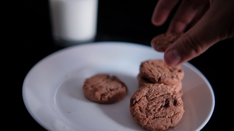 Hand slowly stacking chocolate chip cookies on plate next to blurry glass of milk. Close-up of fresh milk and white plate of tasty cookies with pitch-black background. Sweet food and snacks