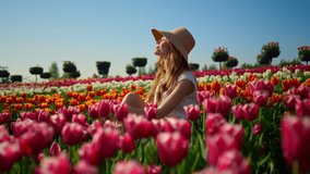 Fancy woman enjoying sun in beautiful holland tulip field. Cheerful woman taking off sunhat in summer sunny day. Relaxed girl turning face to sun in spring flower garden. Girl in blooming spring park.