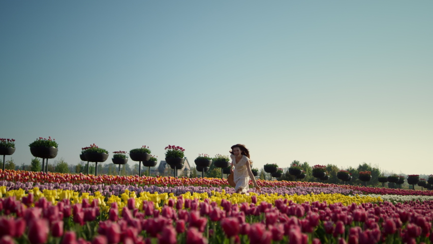 Young woman running through tulip field. Beautiful girl moving fast across spring flowers garden in white dress. Happy girl in straw hat running in tulip garden. View of springtime flower garden. Royalty-Free Stock Footage #1085534507