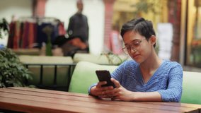 Movement shot of a cheerful Asian Indian young beautiful modern female sitting in an outdoor sidewalk cafe and restaurant smiling while engrossed in a mobile phone or using a smartphone