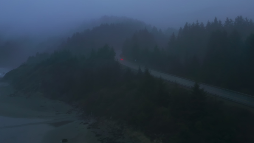 Aerial view of the grayish skyline above the picturesque highway with cars driving along. Dense, lush forest surrounds a coastal road with thick fog above. High quality 4k footage Royalty-Free Stock Footage #1085537105