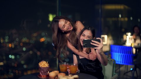 Asian woman friends in meeting and using smartphone selfie together while dinner at skyscraper rooftop restaurant in metropolis at night. Beautiful female enjoy outdoor lifestyle nightlife in the city