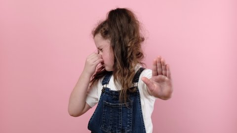 Portrait of little girl kid grabbing nose with fingers, feeling unpleasant smell, shocked with disgusted fart, posing isolated over pink color background wall in studio. People emotion concept