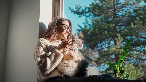 A young adult cheerful woman wearing glasses petting her cute cat while sitting on a windowsill on a sunny day.