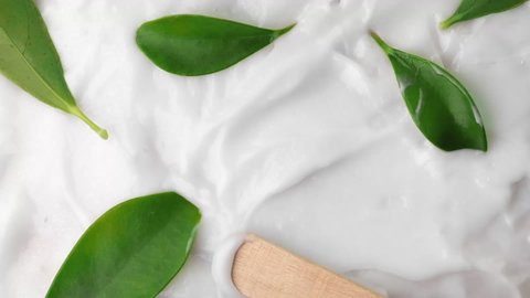 White cream smearing with wooden Spatula. Green fresh natural leaves and Liquid cream, cosmetic texture. Moisturizing balm close-up. Makeup and beauty products. Organic cosmetics, medicine. Top view