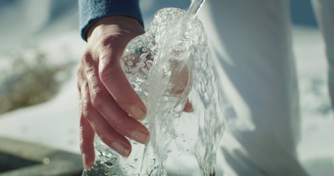 Cinematic macro of thirsty female tourist filling glass bottle with fresh pure cold water from natural spring while hiking in distance during exploration vacation travel in snowy mountains in winter.
