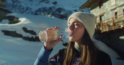 Cinematic shot of thirsty female tourist is drinking fresh pure cold water from glass bottle filled from natural spring while hiking during exploration vacation travel in snowy mountains in winter.