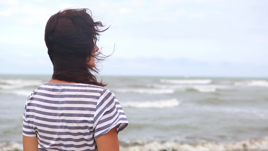 A brunette woman in a striped dress with long hair fluttering in the wind stands by the sea, looks at the waves, holds and removes her hair from her face. Tourism, beach holidays, breeze