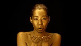 Female With Golden Skin Blowing Gold Dust Looking At Camera Holding Hands Near Face Posing Over Black Studio Background. Beauty And Fashion Art Concept. Slow Motion