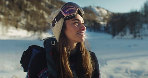 Cinematic shot of carefree young female tourist with backpack is taking off ski goggles and breathes deeply fresh air while hiking on amazing nature landscape of snowy vastness in mountains in winter.