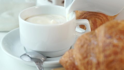 Milk is poured from milk jug into cup of hot cappuccino coffee with creamy foam on background of fresh baked french croissants. Breakfast