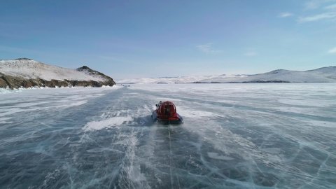 Aerial view on the hovercraft driving on cracked snowy ice of Baikal along the rocky cliff. Drone follows the vehicle. Beautiful winter landscape of lake Baikal
