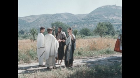 1960s: People in period costume talking on road. Woman with clapboard. Guards approach group on road.