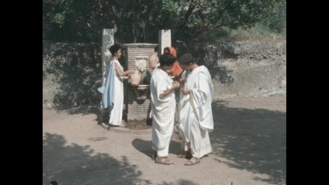 1960s: People in period costume, women getting water from fountain, men talking in foreground.