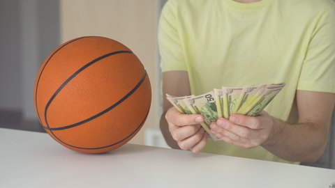 Lucky man with basketball ball counting dollar banknotes. Close up of male hands count money cash payout after win at sports betting. Concept of betting, gambling.