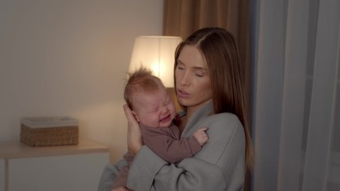 Worried Young Mother Holding Crying Newborn Baby In Arms, Caring Millennial Mom Embracing And Soothing Her Upset Infant Son Or Daughter While Standing In Bedroom At Home, Slow Motion Footage