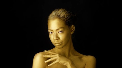 Attractive Lady With Golden Skin Touching Neckline Stroking Body Covered With Golden Paint Posing Over Black Studio Background, Looking At Camera. Makeup And Fashion Art, Beauty Portrait