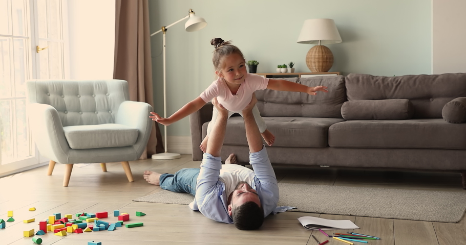 Cute little girl spread arms like wings enjoy freedom in imaginary flight balance in air on strong dad hands. Loving daddy lie on floor with underheating help small daughter imitate plane raise kid up Royalty-Free Stock Footage #1085566922