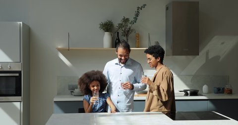 Young African couple and cute 10s daughter standing in kitchen holding glasses, drinking still natural water, reducing thirst in summer day. Healthy lifestyle, home water purification system concept