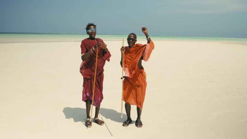 Two Young Happy Masai Tribe Members in Traditional Red Dresses and Sunglasses Dancing on the Sand Beach of the Indian Ocean. Zanzibar. Tanzania. 4K 50fps Royalty-Free Stock Footage #1085569976