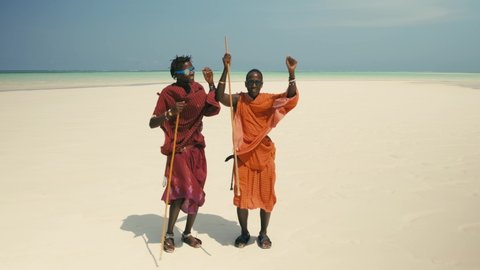Two Young Happy Masai Tribe Members in Traditional Red Dresses and Sunglasses Dancing on the Sand Beach of the Indian Ocean. Zanzibar. Tanzania. 4K 50fps