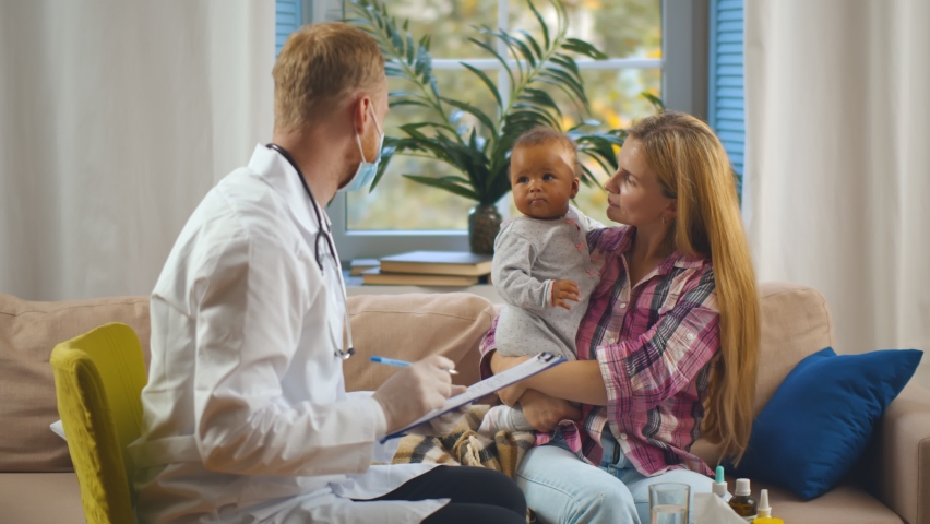 Caucasian woman with adopted mixed race baby consulting pediatrician at home. Young doctor in safety mask visiting mother with kid at home. Healthcare and adoption concept