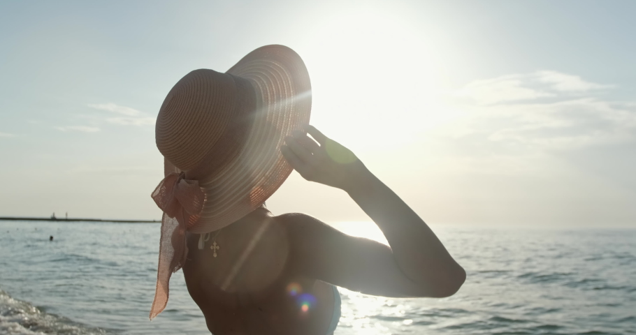 Slim woman in bikini swimsuit and pink hat with ribbon bow enjoys view of blue sea water at warm sunlight on beach slow motion | Shutterstock HD Video #1085576237
