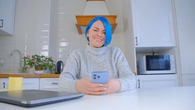 Happy white woman with blue hair using mobile phone. Cute Caucasian female with dyed hair typing message on modern smartphone with triple camera. Communicative person messaging on social media app