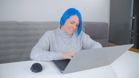 Happy millenial woman typing on computer. Stylish white female with dyed blue hair working distant from home on lockdown. Beautiful Caucasian person doing distant work online on notebook pc
